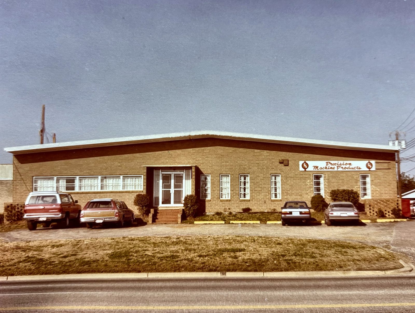 Color photo of a tan brick Precision Machine Products building with 1980s cars